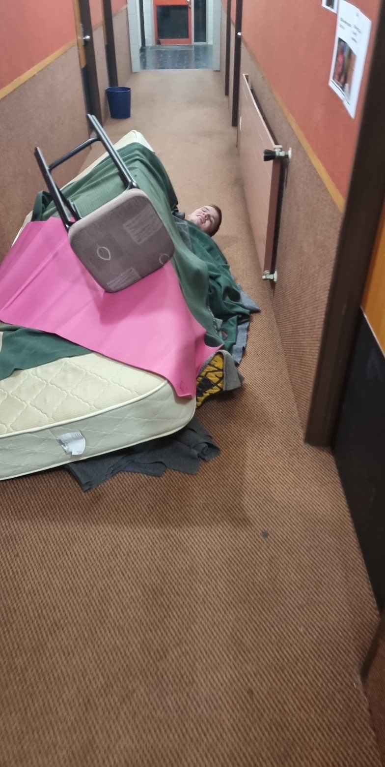 Me asleep on a barracks corridor floor under a mattress, a chair, two blankets and a pink exercise mat